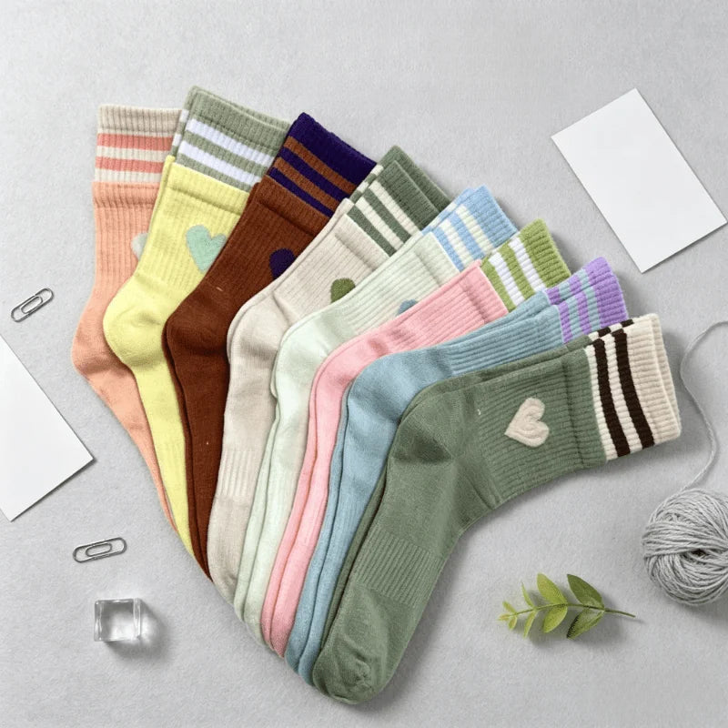 A set of ten pastel-colored crew socks arranged in a fan shape on a light gray surface, styled with paper clips, a small card, a glass cube, a green leaf sprig, and a ball of gray yarn.
