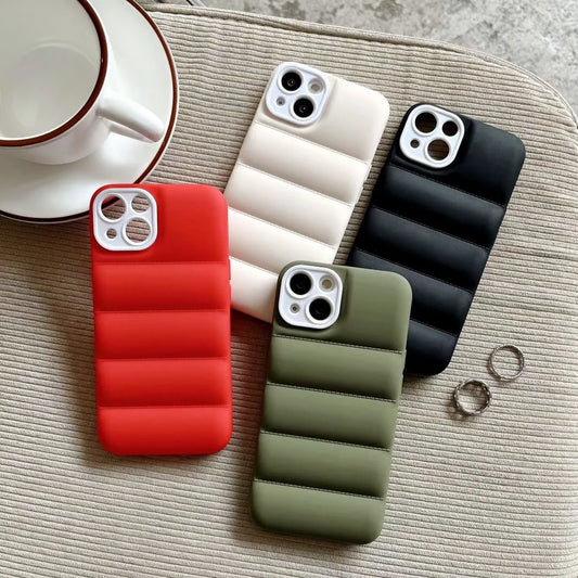 Four quilted phone cases in red, cream, black, and olive green are arranged on a textured beige fabric surface next to a white teacup and saucer, with two silver rings nearby.
