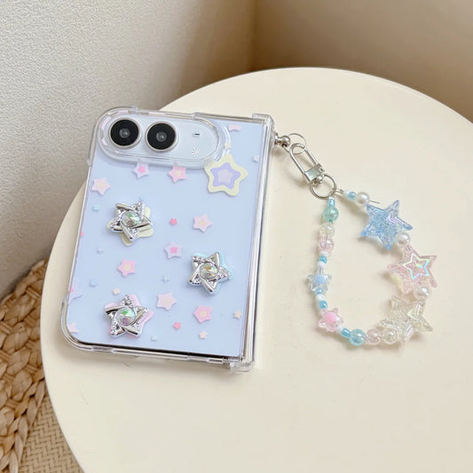 A clear phone case with a pastel light blue back, decorated with iridescent star charms, small pastel star stickers, and a matching beaded star strap, shown resting on a cream-colored surface.