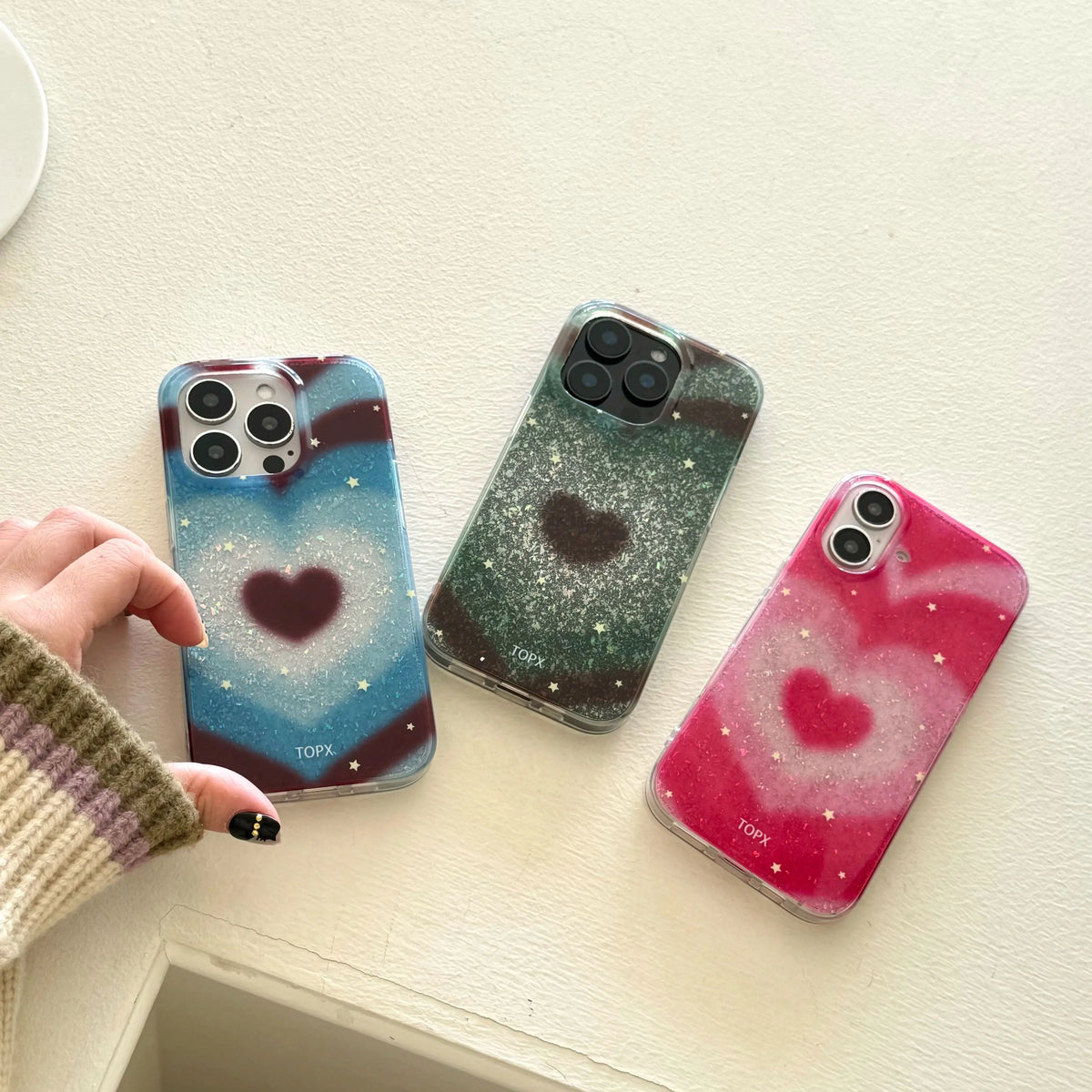 Three phone cases with heart designs are displayed on a light beige surface. A hand wearing a beige and purple striped sweater is holding the leftmost case.