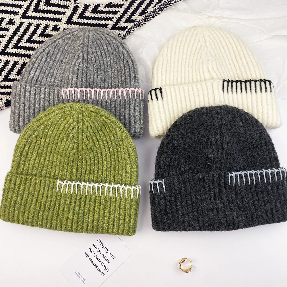 Four ribbed knit beanie hats arranged in a flat lay on a white surface with a black-and-white geometric textile in the top left. The beanies are in gray, cream, olive green, and charcoal black, each with contrasting stitched decorative details in light pink, black, white, and light blue respectively.