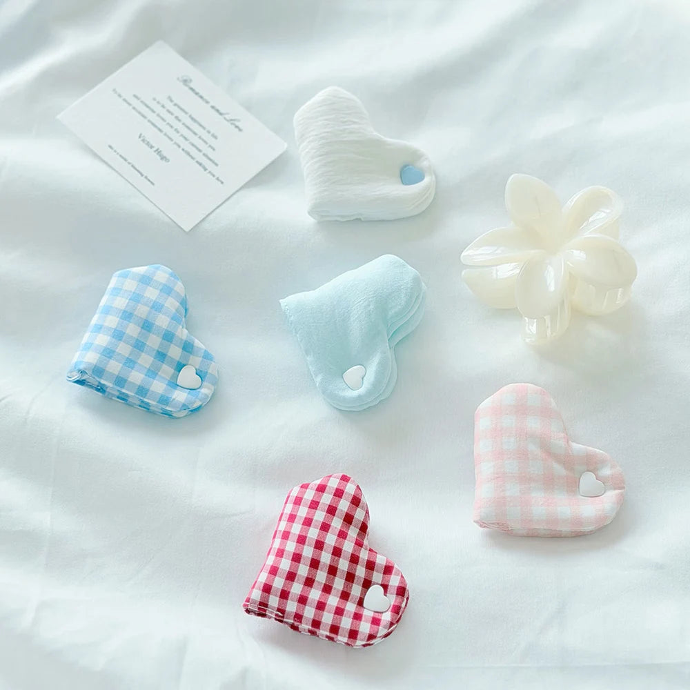 Six heart-shaped hair claw clips in various colors and textures are arranged on a white fabric background. The clips include different patterns: two are light blue gingham, one is solid light blue, one is solid cream, one is red gingham, and one is pink gingham.