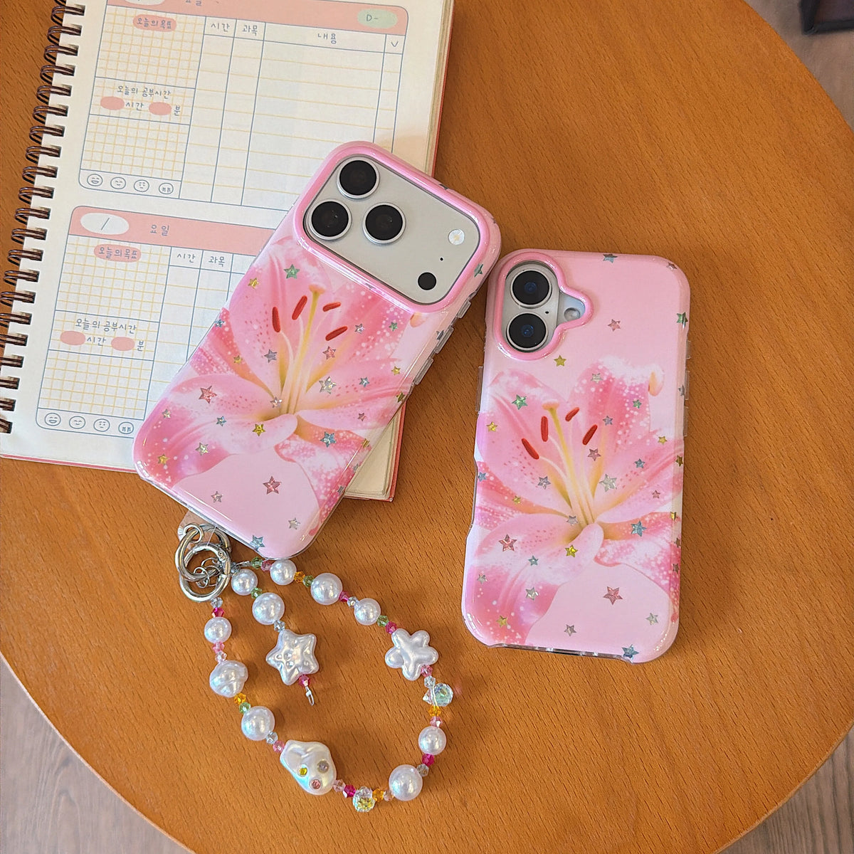Two pink floral phone cases with a matching pearl charm strap, styled on a wooden table next to a planner notebook.