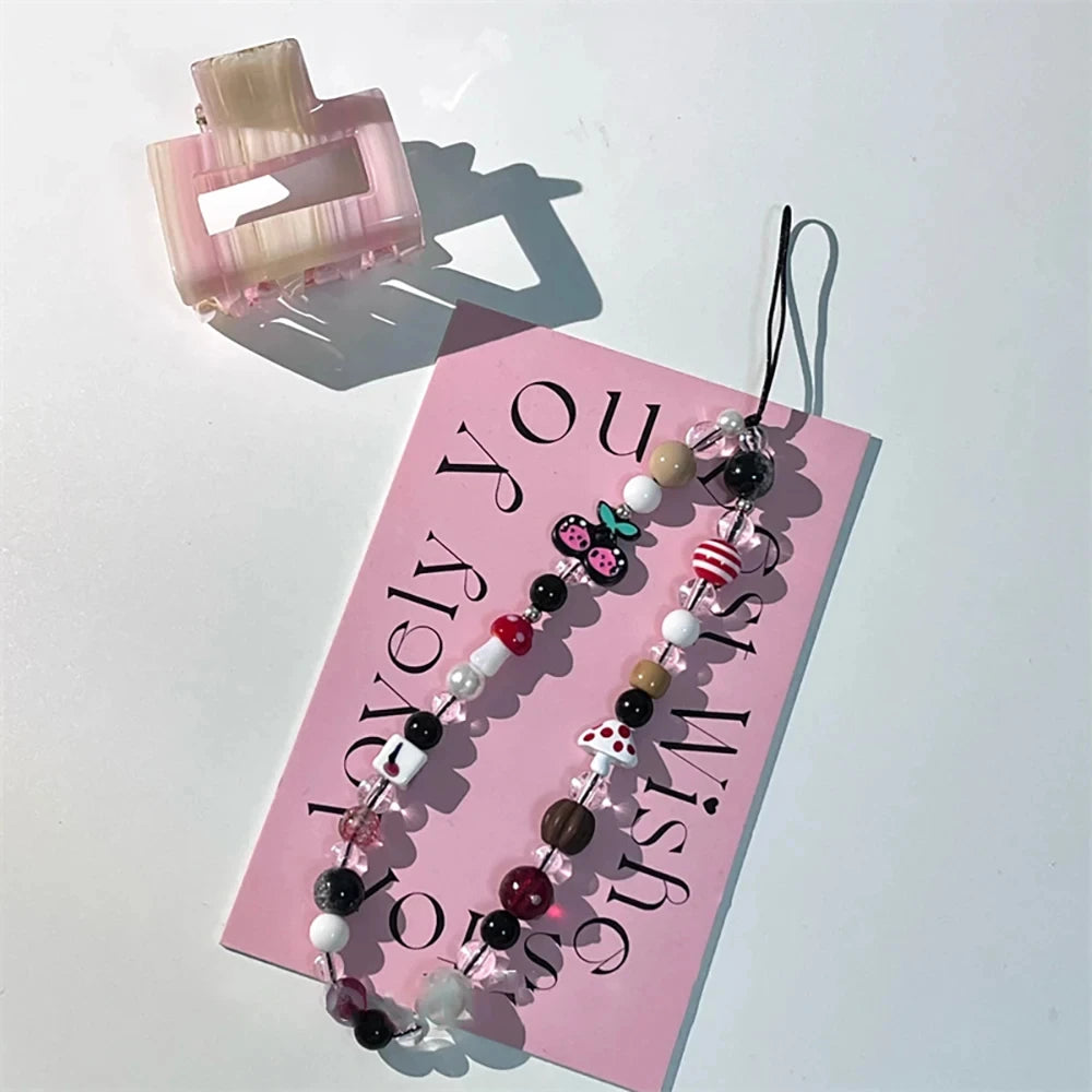 A pink beaded phone strap with a black cord and decorative charms, displayed on a pink card with a translucent pink hair claw clip on a white background.