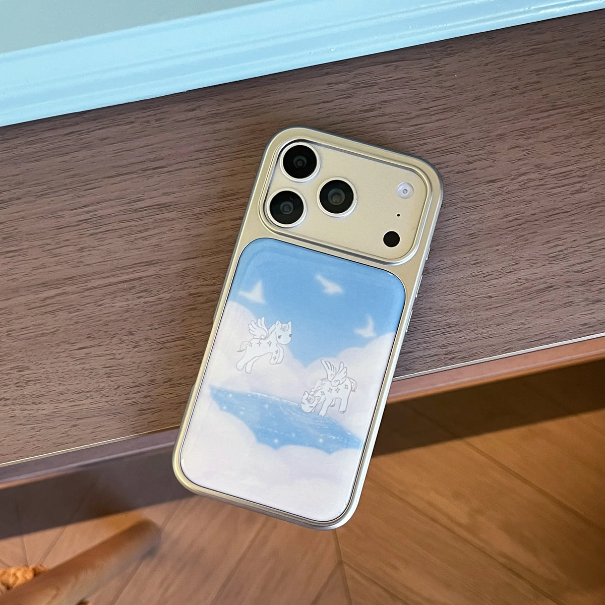 A gold-colored phone case with a printed sky and cloud design, shown resting on a wooden table.