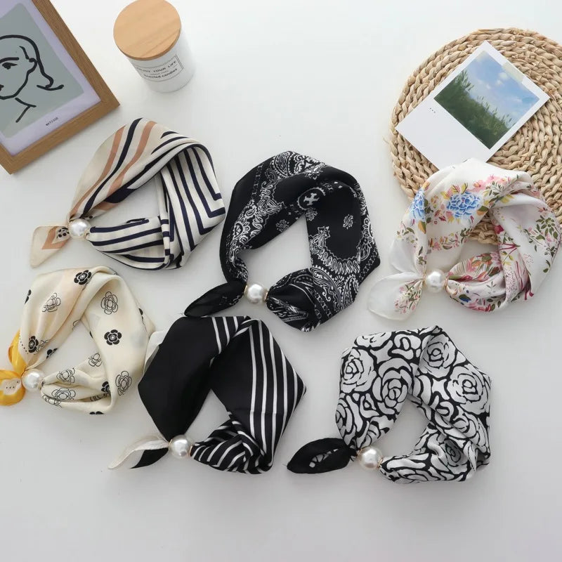 Five silk hair scrunchies with pearl accents are arranged in a flat-lay on a white surface. The scrunchies feature various patterns including stripes, florals, and roses in black, white, beige, and pastel colors.