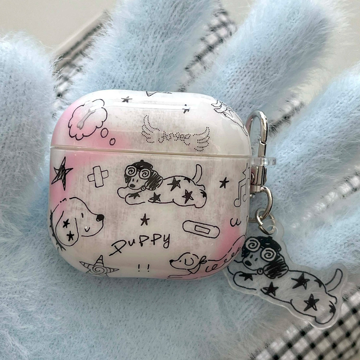 A white and pink AirPods case decorated with black doodle illustrations of puppies, stars, and musical notes, featuring a clear acrylic keychain charm with a matching puppy design and a silver clasp, resting on a soft light blue fuzzy blanket.