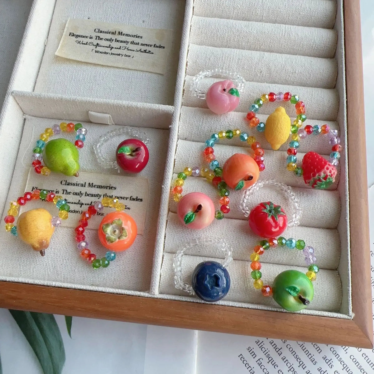A set of colorful beaded fruit-themed rings displayed inside a light wood jewelry box with cream lining. The rings feature small multicolored beads and large fruit charms including a pear, apple, strawberry, orange, blueberry, and lemon.