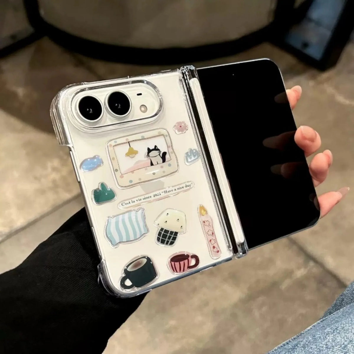 A clear TPU protective case for a folding smartphone, decorated with cute pastel cartoon illustrations of pillows, cups, and a cat, held open in a hand.