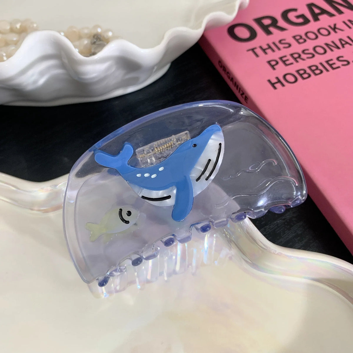A translucent clear hair claw clip featuring a blue whale and small fish design, styled on a white surface next to a pink book.