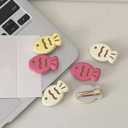 Five fish-shaped hair clips in pastel yellow, pink, and cream are arranged on a light surface next to a closed silver laptop. One cream clip shows its rose-gold metal clasp.