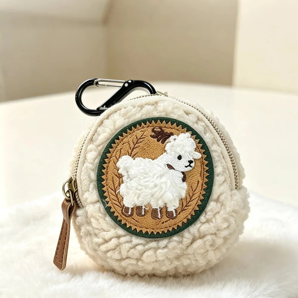 A round cream-colored sherpa coin purse with a black carabiner clip and a brown zipper pull, featuring a circular embroidered sheep patch on the front.