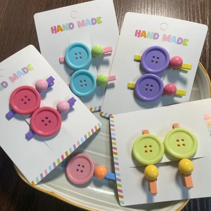 Four colorful button-shaped hair clips on white 'HAND MADE' cards, arranged on a tray over a dark wood surface. The clips feature pastel shades of pink, purple, blue, green, yellow, and orange.