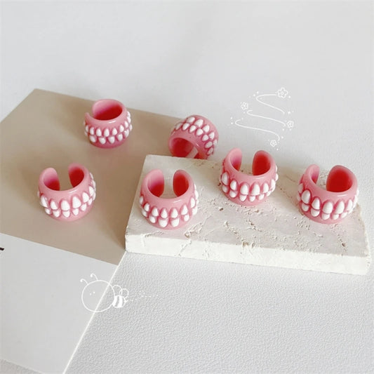 Five pink silicone ring-shaped accessories with white tooth-like details, arranged on a white surface with a beige rectangular block. No person or real environment is present, so this is Product/Packaging rather than Lifestyle.