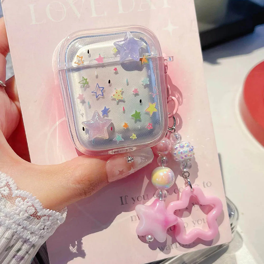 A hand holds a clear plastic AirPods case decorated with pastel star and galaxy designs, attached to a pink star-shaped charm and iridescent beads, against a pink card background.