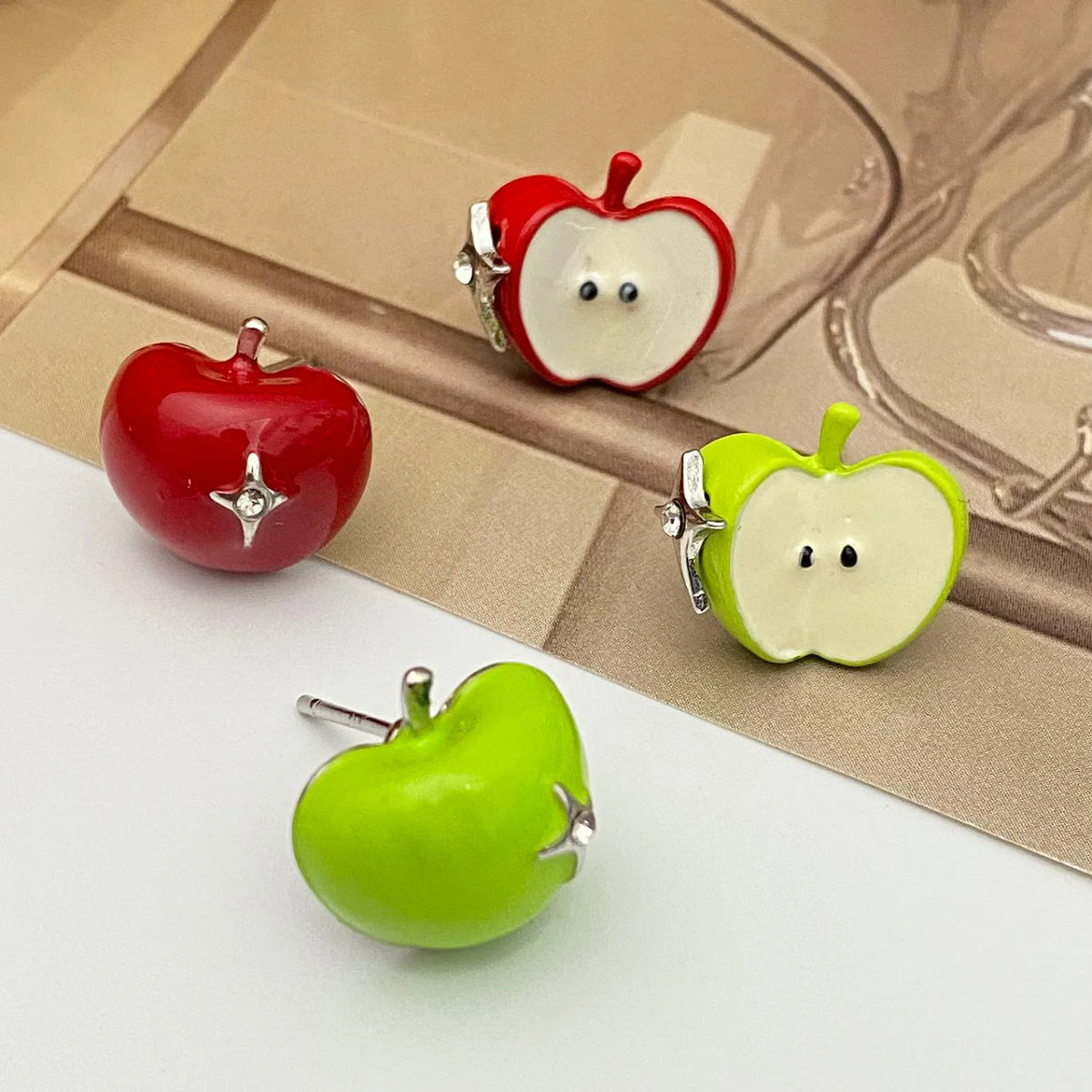 Three glossy apple-shaped stud earrings displayed on a split white and beige surface. One earring is a solid red apple, one is a red apple half with white flesh and small black eyes, and one is a green apple half with white flesh and small black eyes.