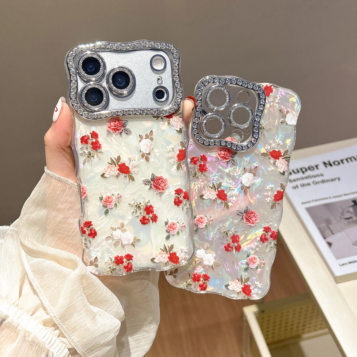 A hand holds two decorative phone cases with floral embellishments and rhinestone-embellished camera areas, shown against an indoor room background.