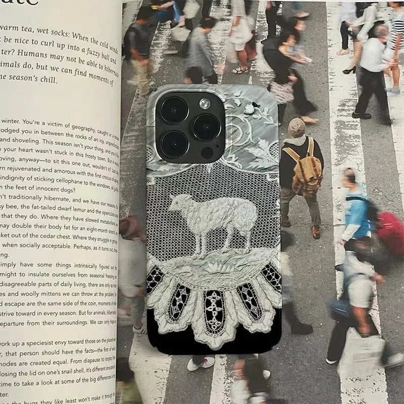 A black mesh phone case with white embroidered lace details and a white embroidered monogram pattern, shown on a smartphone. The phone case is resting on an open book with a blurred New York City street scene printed on the page behind it.