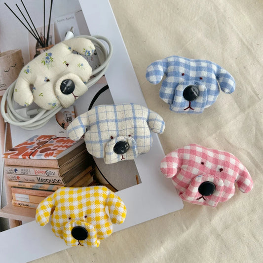 Four small plush dog-shaped cable organisers in different gingham colorways are arranged on a light fabric surface with books and a white tray as styled props. The dog faces are cream, light blue, pink, and yellow, each with black stitched eyes and nose details.