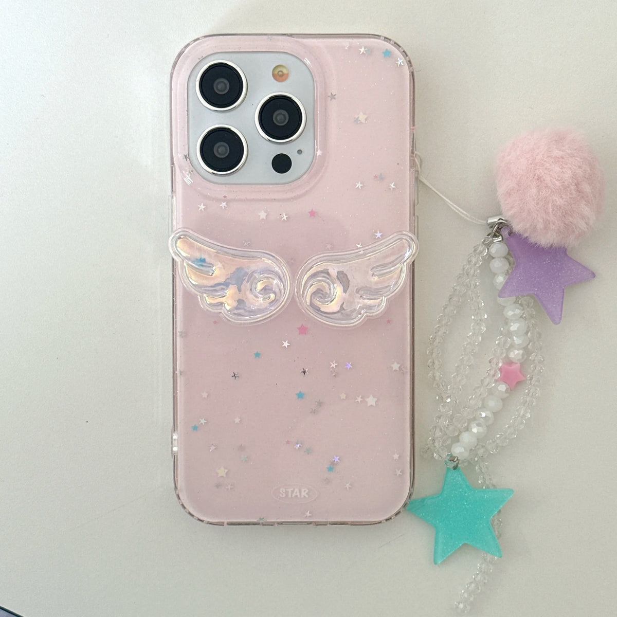 A light pink phone case with clear glitter and small star details, featuring a 3D clear angel wing design on the back. The case is shown next to a decorative charm with a pink pom-pom, a purple star, a teal star, and white beads on a plain light background.