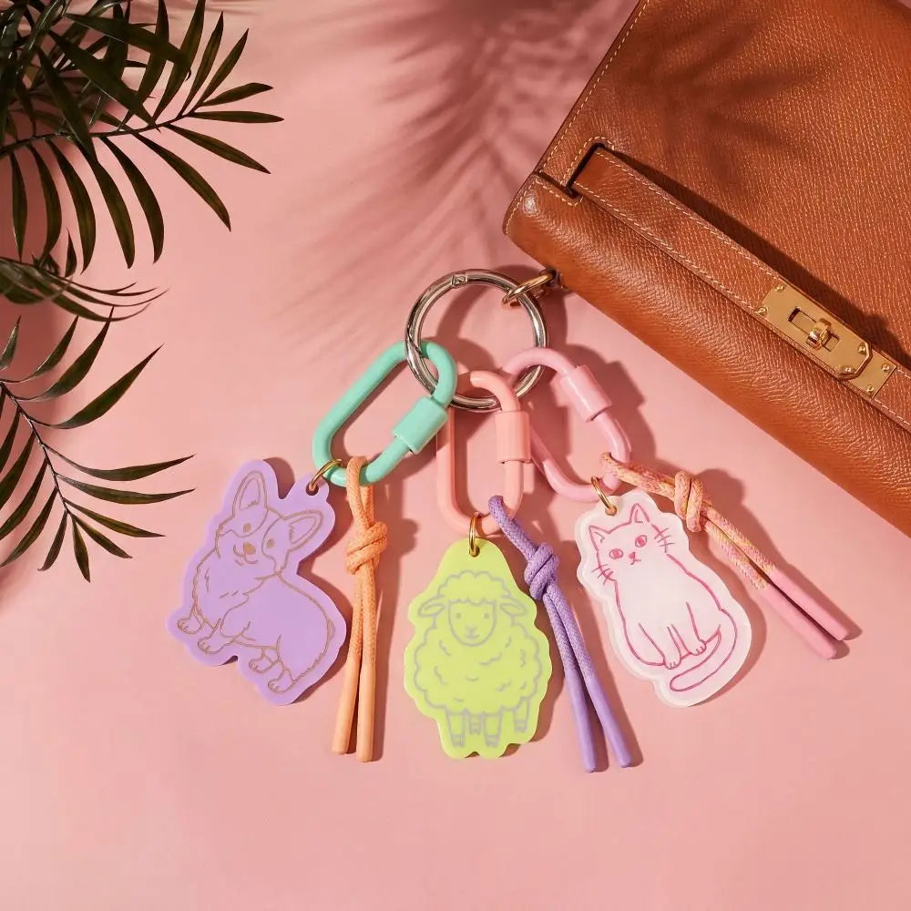 A set of four colorful animal-shaped keychains with rope ties and metal carabiners, displayed on a pink background next to a brown leather bag and green palm leaves.