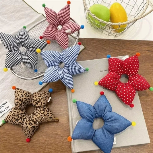 Six fabric flower-shaped hair bows in different patterns (black-and-white gingham, pink-and-white gingham, blue-and-white gingham, red-and-white polka dots, leopard print, and denim blue) arranged on a wooden table with books and a wire fruit basket in the background.