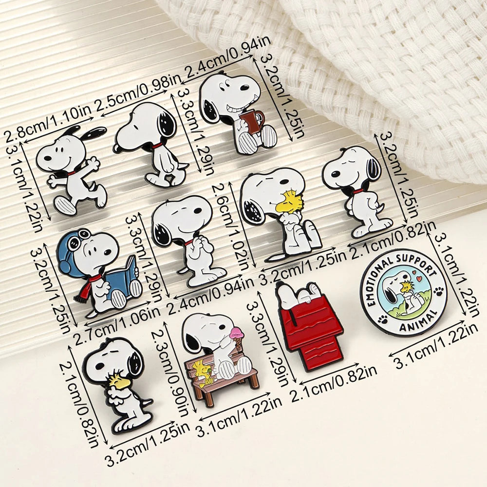 A set of Snoopy enamel pins arranged on a light beige background with printed dimensions in centimeters and inches. The pins include Snoopy in various poses, Snoopy reading, Snoopy with a yellow bird, Snoopy in a red hat, Snoopy in a red doghouse, and a circular pin reading 'Emotional Support Animal'.