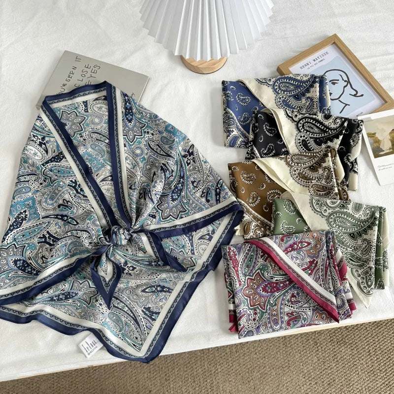 A styled flat-lay of multiple folded and tied silk scarves with intricate paisley patterns in various colors, arranged on a white textured fabric surface alongside a pleated lamp, a framed print, and a book.