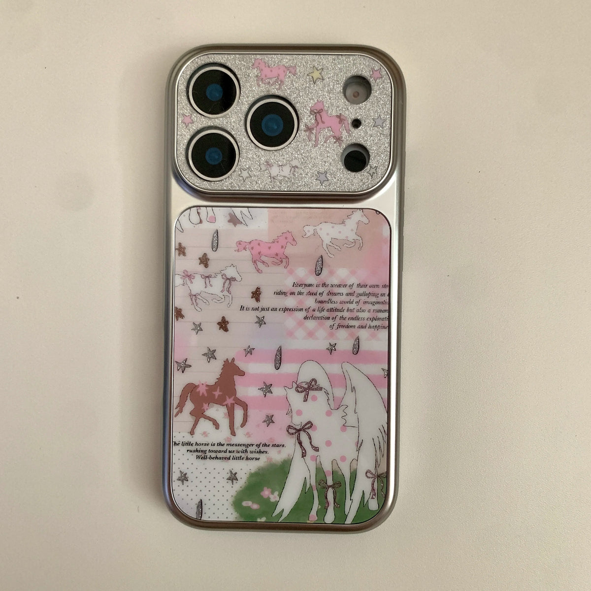 A silver iPhone case featuring a whimsical unicorn illustration design with pink, brown, and white horses, stars, and text on a light beige background.