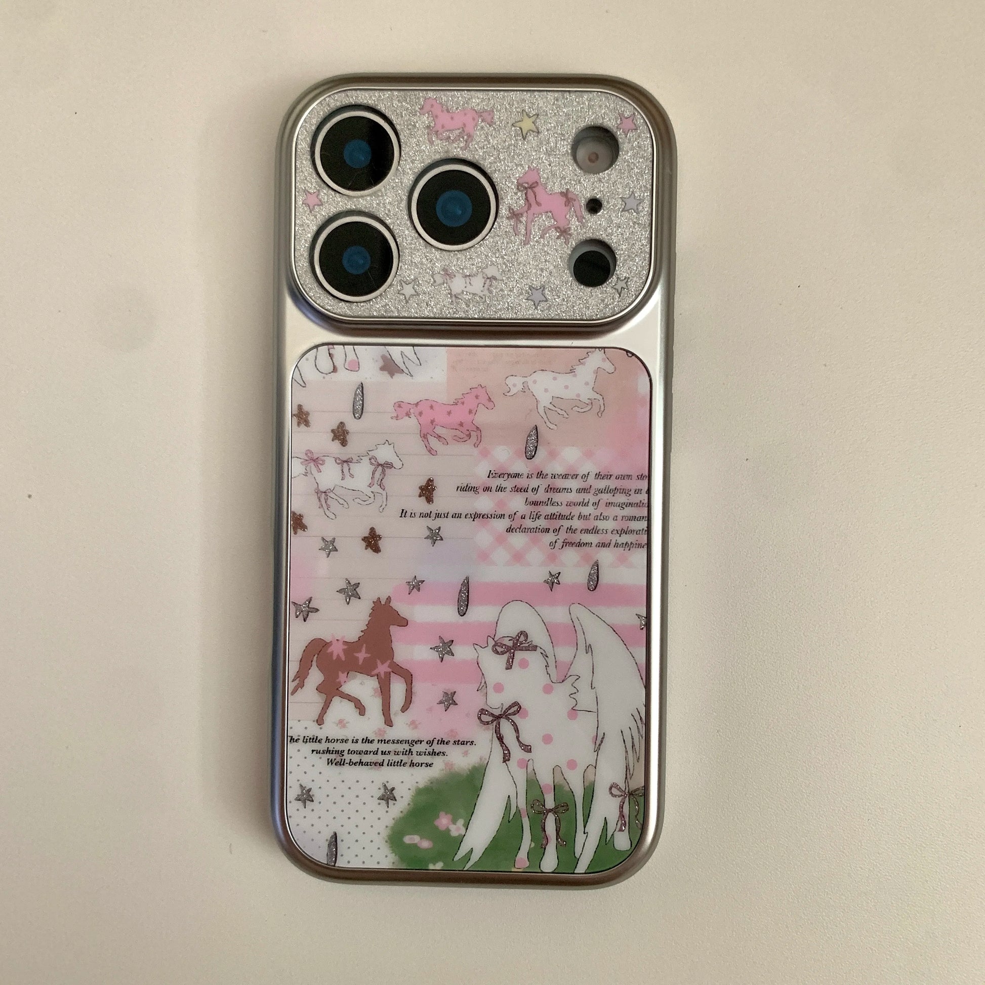 A silver iPhone case featuring a whimsical unicorn illustration design with pink, brown, and white horses, stars, and text on a light beige background.
