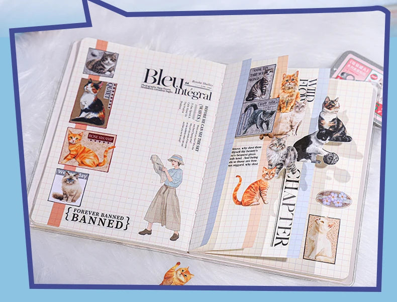 An open handmade scrapbook journal with cat-themed collage pages, featuring illustrations of various cats, vintage-style text, and decorative elements, set against a light blue background.