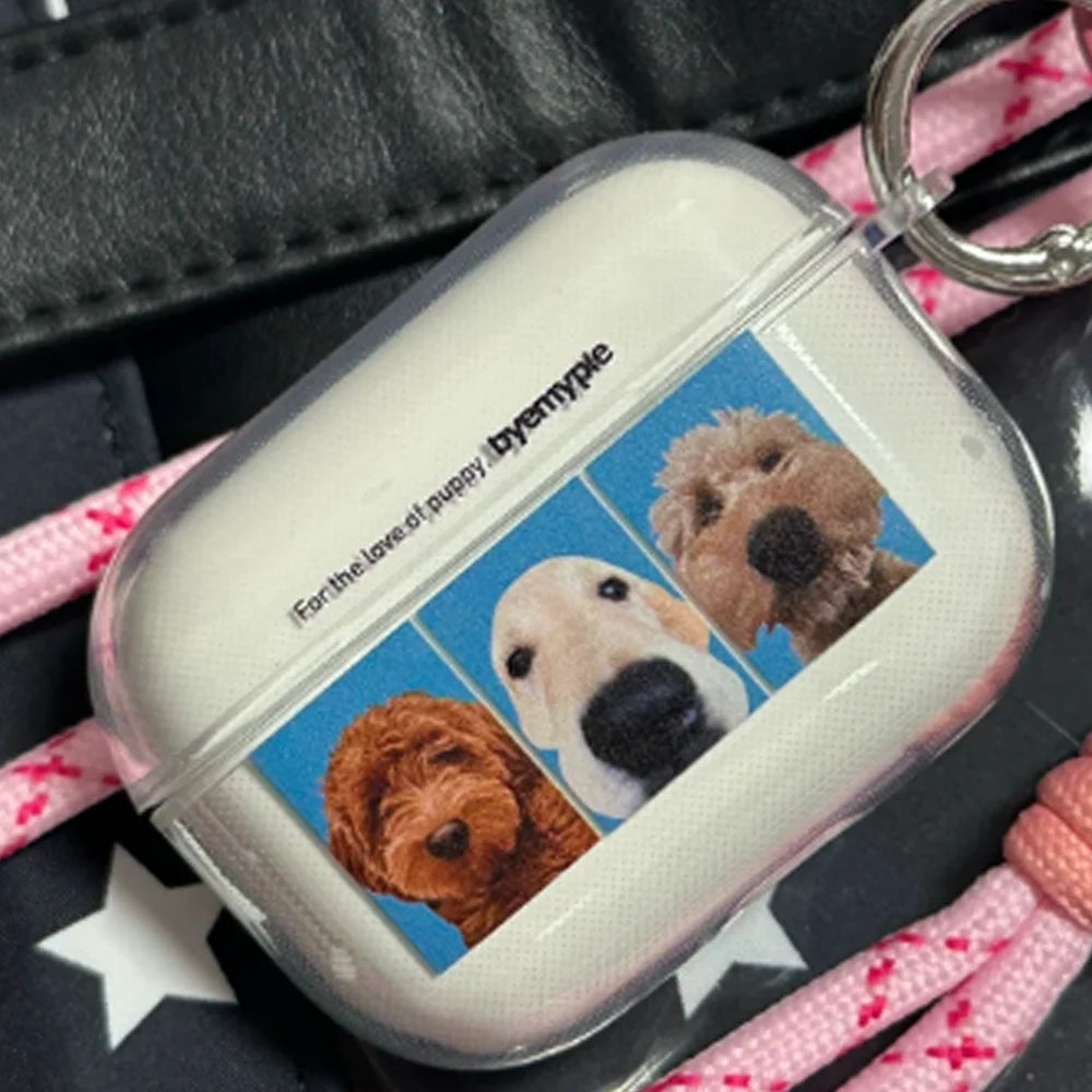 A clear AirPods case with a printed photo of three dogs on a blue background and the text 'For the love of puppy byenypyple', shown resting on a black leather surface with a pink patterned strap.