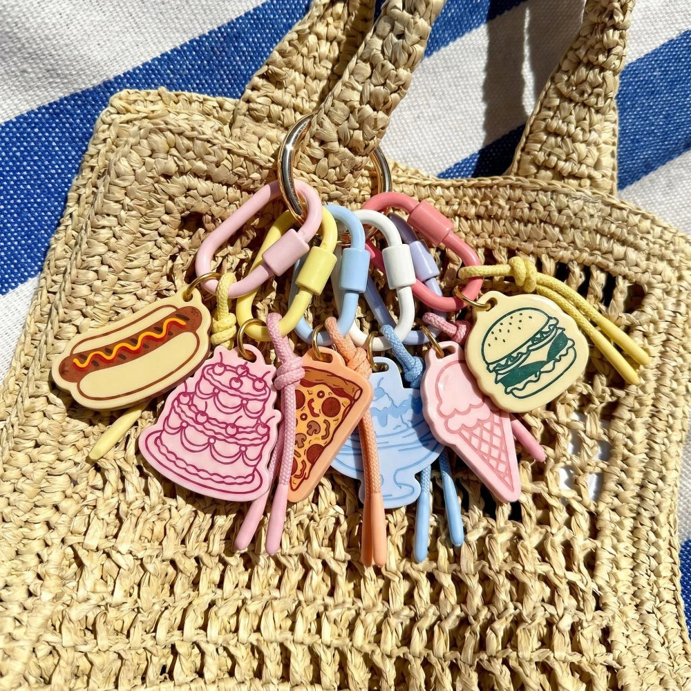 A set of colorful silicone food-themed keychain charms, including a hot dog, cake, pizza slice, blue straw, ice cream cone, and hamburger, attached to a metal key ring and draped over a woven straw tote bag.