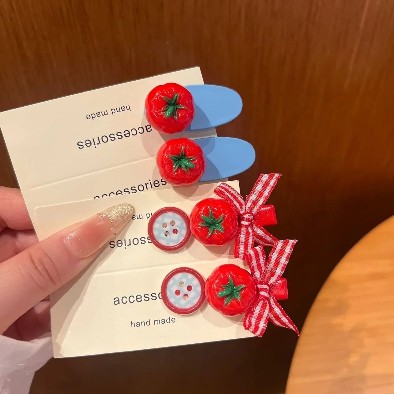 A hand holds three small accessory cards featuring red tomato-shaped clips with green stems and two red-and-white gingham ribbon bows with matching decorative buttons, against a warm brown wooden background.