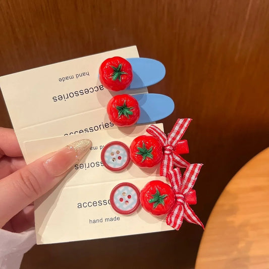 A hand holds three small accessory cards featuring red tomato-shaped clips with green stems and two red-and-white gingham ribbon bows with matching decorative buttons, against a warm brown wooden background.