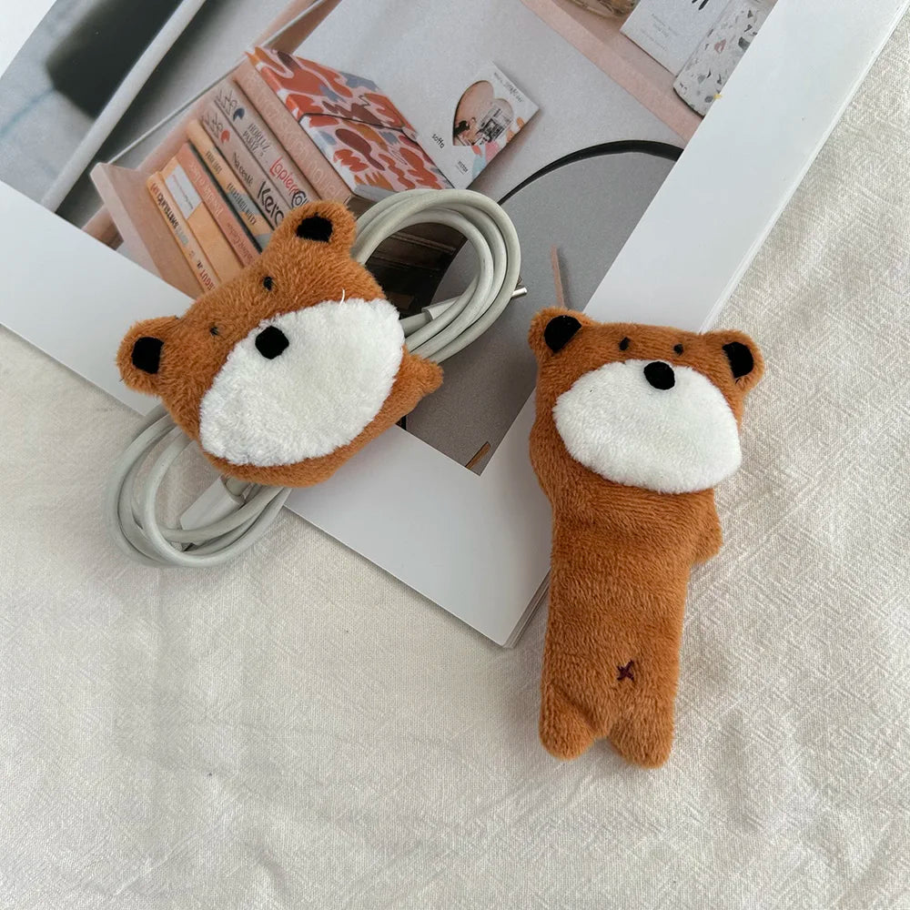 Two plush fox-shaped cable clips in brown and white, shown on a light fabric surface with a white tray and books in the background.