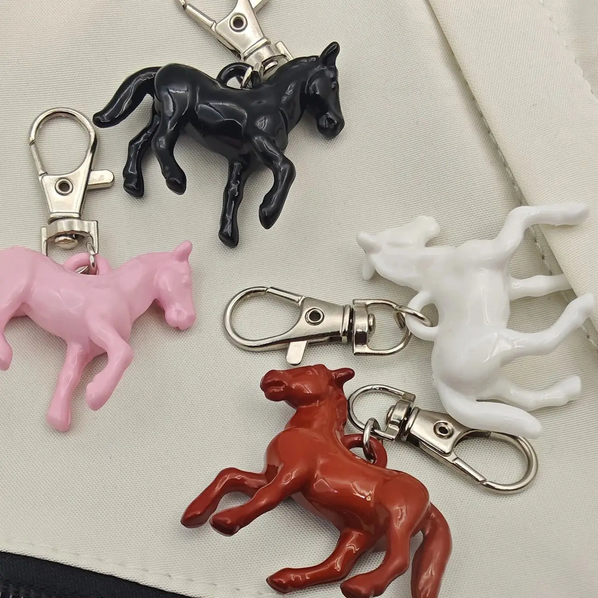 Four small horse-shaped keychains in pink, black, white, and reddish brown, each attached to a silver lobster clasp, arranged on a light fabric background.