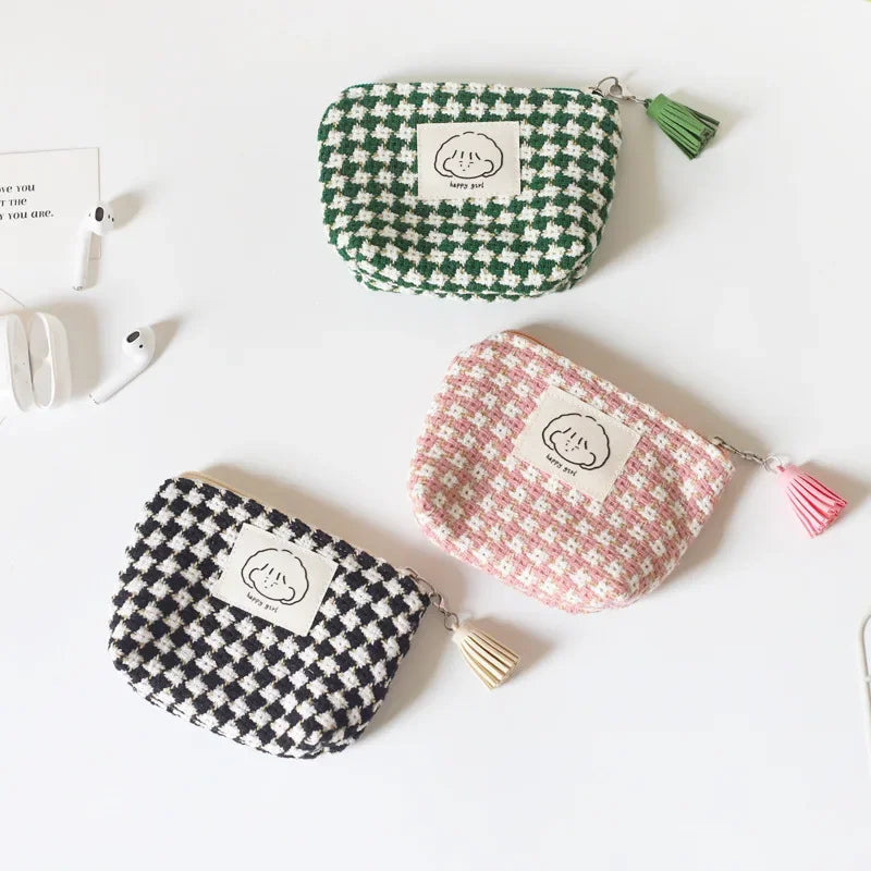Three small woven zip pouches with tassel charms are arranged on a white surface. One pouch is green and white houndstooth, one is pink and white houndstooth, and one is black and white houndstooth.