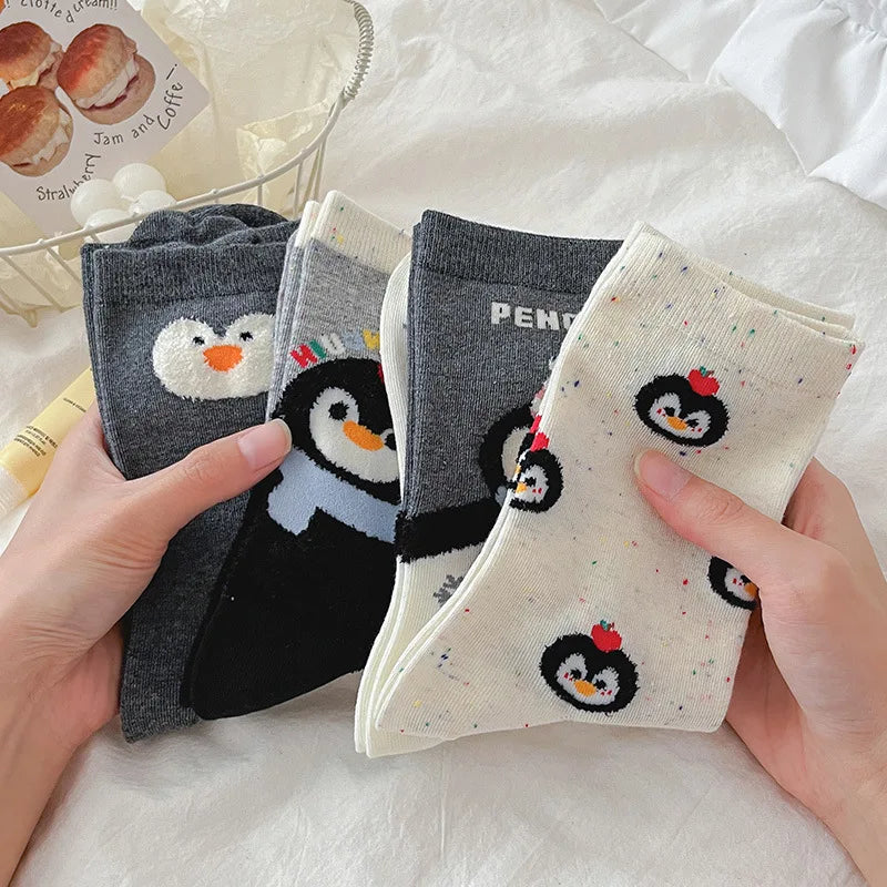Four pairs of socks held in two hands against a white fabric background. The socks feature penguin designs: two dark gray pairs with black-and-white penguin faces and one light gray pair with a black-and-white penguin body, plus one cream pair with a small black-and-white penguin and multicolor speckles.