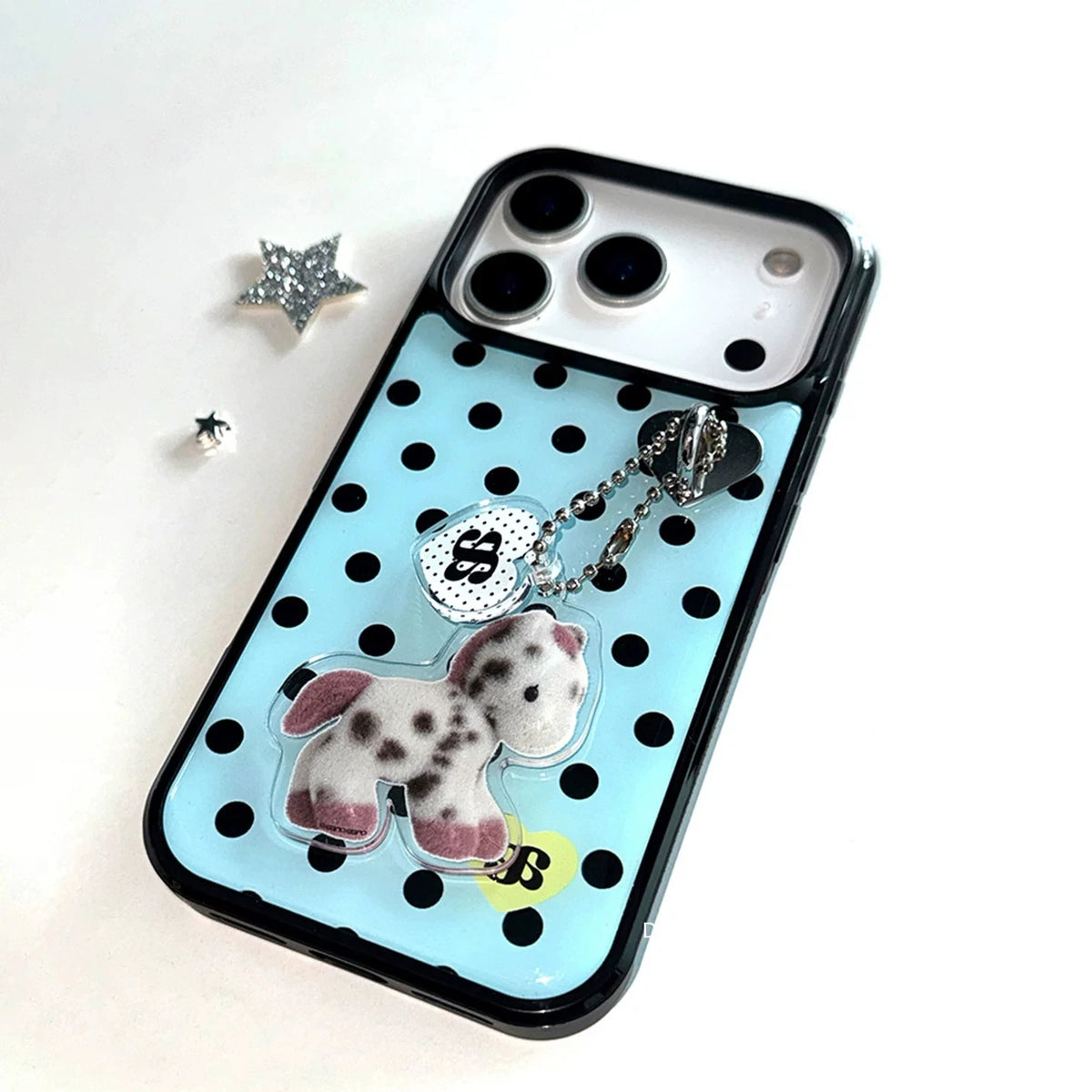 A light blue phone case with black polka dots, featuring a charm with a dalmatian dog figure, a silver heart charm, and a small yellow accent, displayed on a white background.