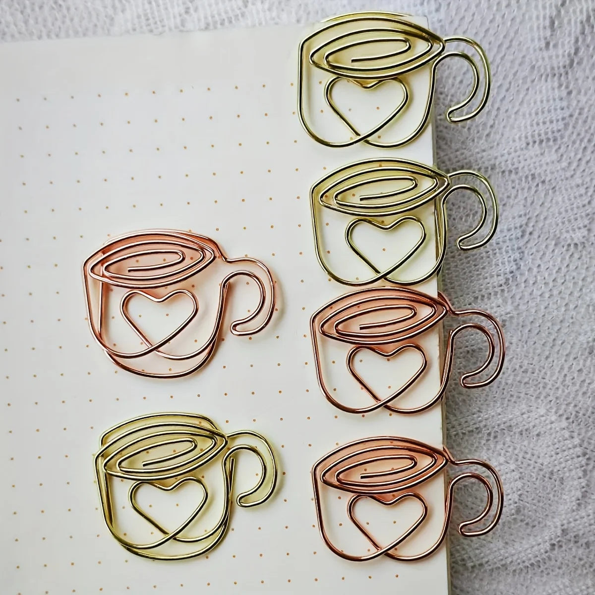 Four metallic paperclip bookmarks shaped like coffee cups with heart cutouts, shown in gold and rose gold finishes on a white dotted notebook page with a textured white fabric background.