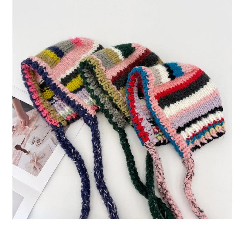 Three colorful striped knit winter hats with long ear flaps and braided ties, arranged on a white surface with a small photo card partially visible.