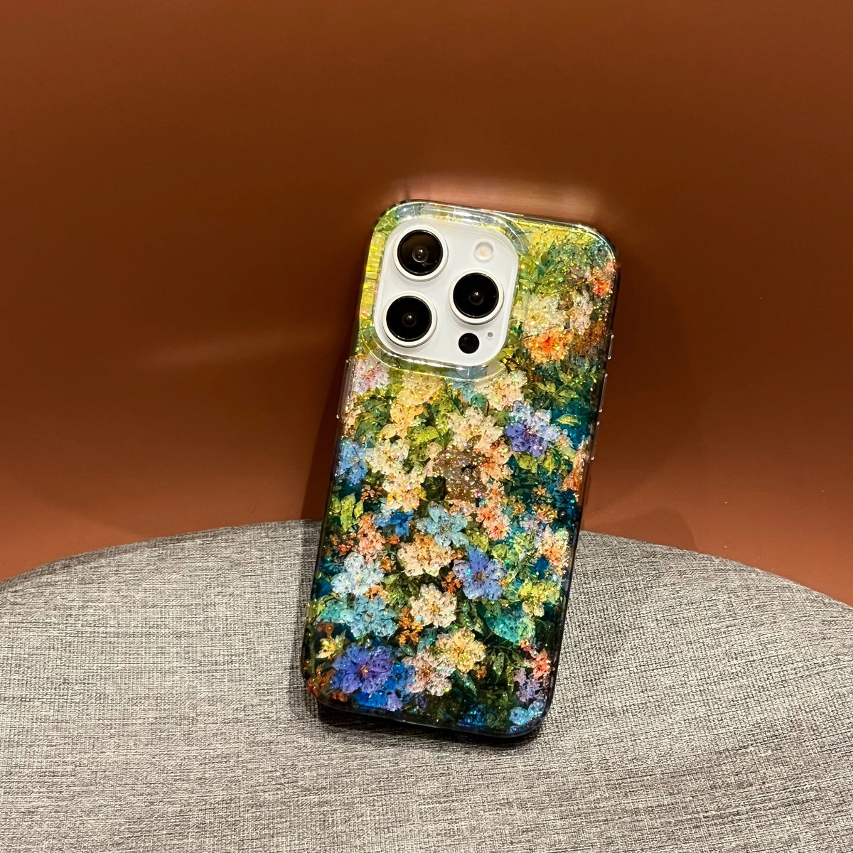 A phone case with a dense, colorful floral pattern in shades of blue, yellow, orange, and green, shown on a smartphone resting on a gray textured surface against a warm brown background.
