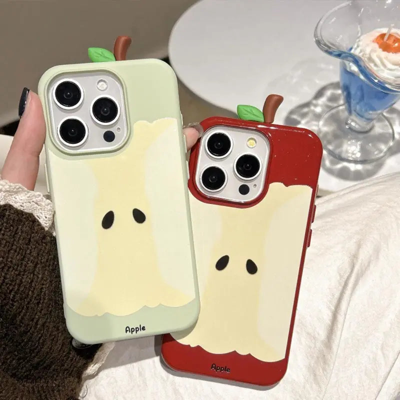 Two Apple iPhone cases featuring a cute ghost-like apple design, held in a hand. One case is a pale sage green with a cream-colored apple slice graphic, and the other is red with a matching cream graphic.
