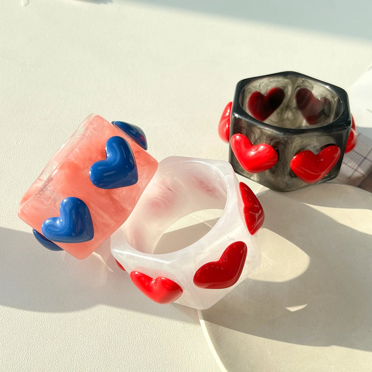 Three translucent resin bangles with raised heart details, displayed on a light surface in bright natural light. One ring is peach-pink with blue hearts, one is white with red hearts, and one is smoky gray with red hearts.
