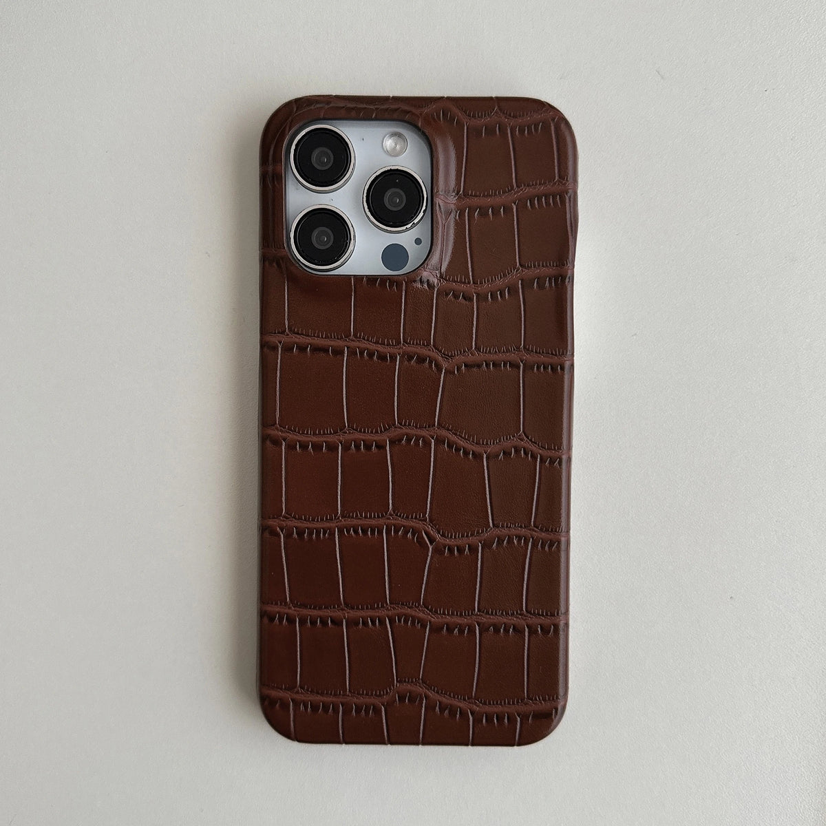 A dark brown crocodile-embossed leather phone case on a silver iPhone, shown from the back against a light gray background.
