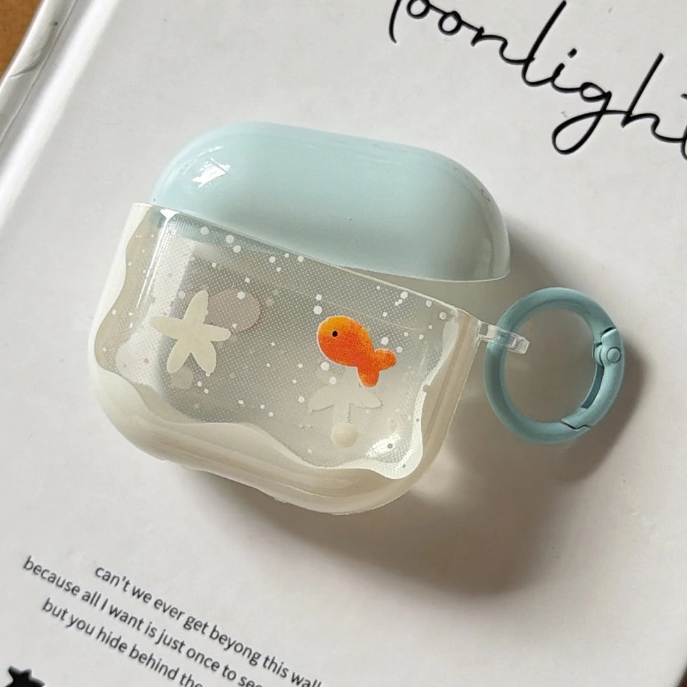A translucent silicone AirPods case with a light blue lid and matching ring clip, decorated with a small orange goldfish and white starfish on a glittery background, resting on a white book page.