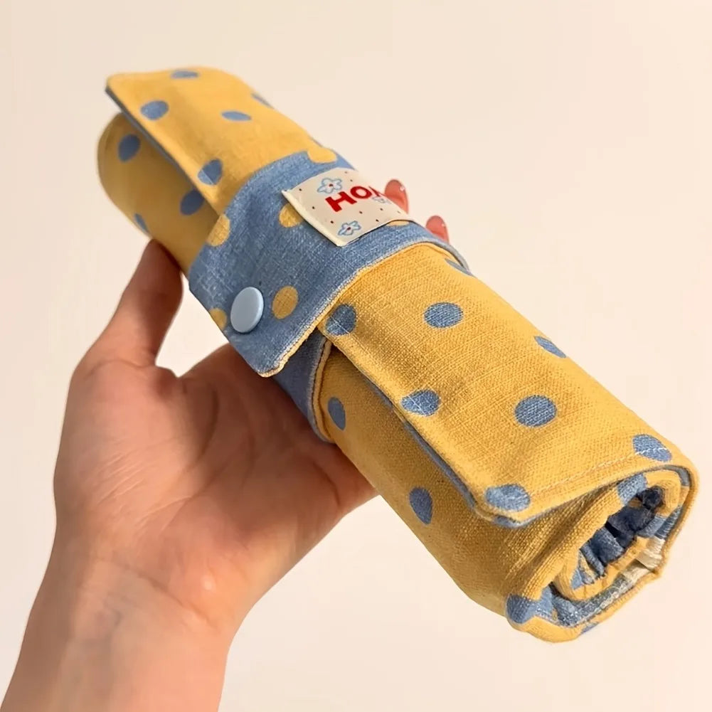 A hand holding a rolled yellow fabric pouch with blue polka dots and a blue strap closure. A small fabric label is visible on the front.
