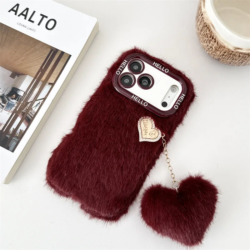 A plush faux-fur phone case in deep burgundy red, featuring a matching heart-shaped fur charm attached by a gold chain. The case is styled on a white surface next to a book and a small woven cup.
