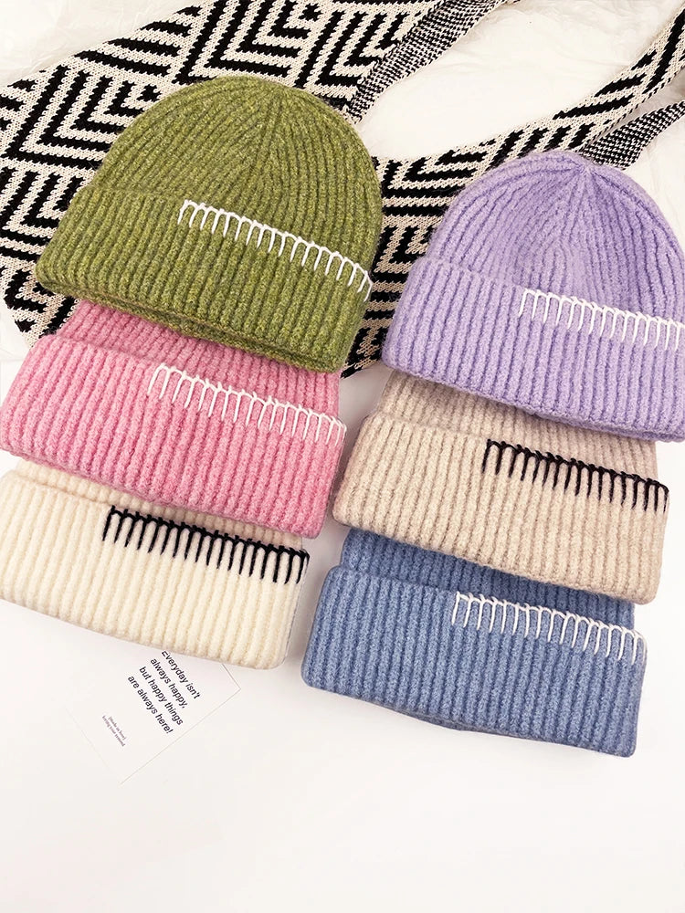 Four ribbed knit beanie hats arranged in two columns on a white surface with a black-and-white patterned textile in the background. The hats are olive green, pink, cream, and lavender on the left, and cream, beige, and dusty blue on the right.